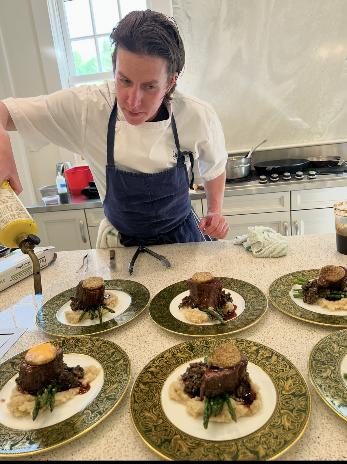 A chef in a white shirt and navy apron platters a dish on a kitchen counter, with several plated meals arranged in front. The meals include a piece of beef, mashed potatoes, asparagus, and a fried egg on decorative green and gold plates.