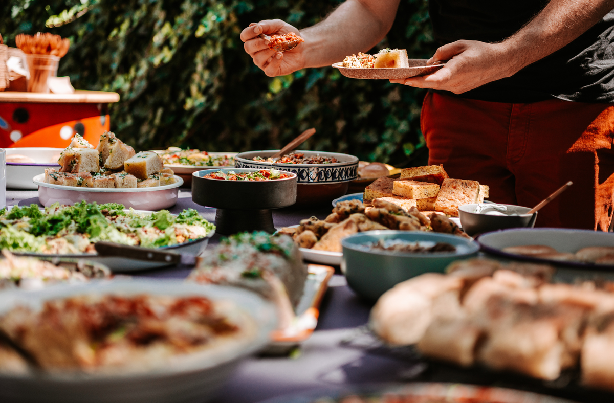 A table laden with an outdoor barbecue spread of breads, salads, and dips, with a person in a black shirt and rust-red pants holding a plate in the background.